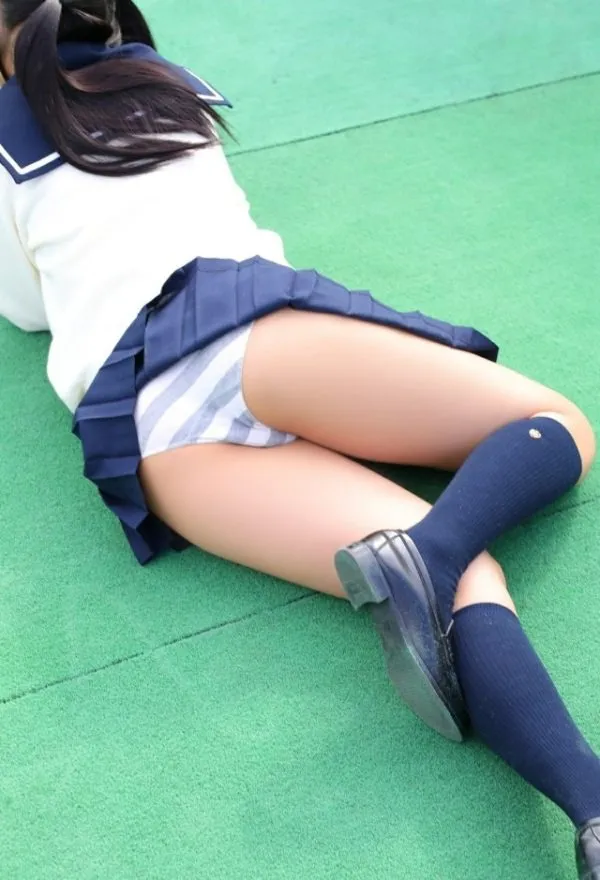 JKが屋外の緑の床にうつ伏せになり、紺色のプリーツスカートをめくって白とグレーのボーダー柄パンツを見せるムチムチ尻の写真