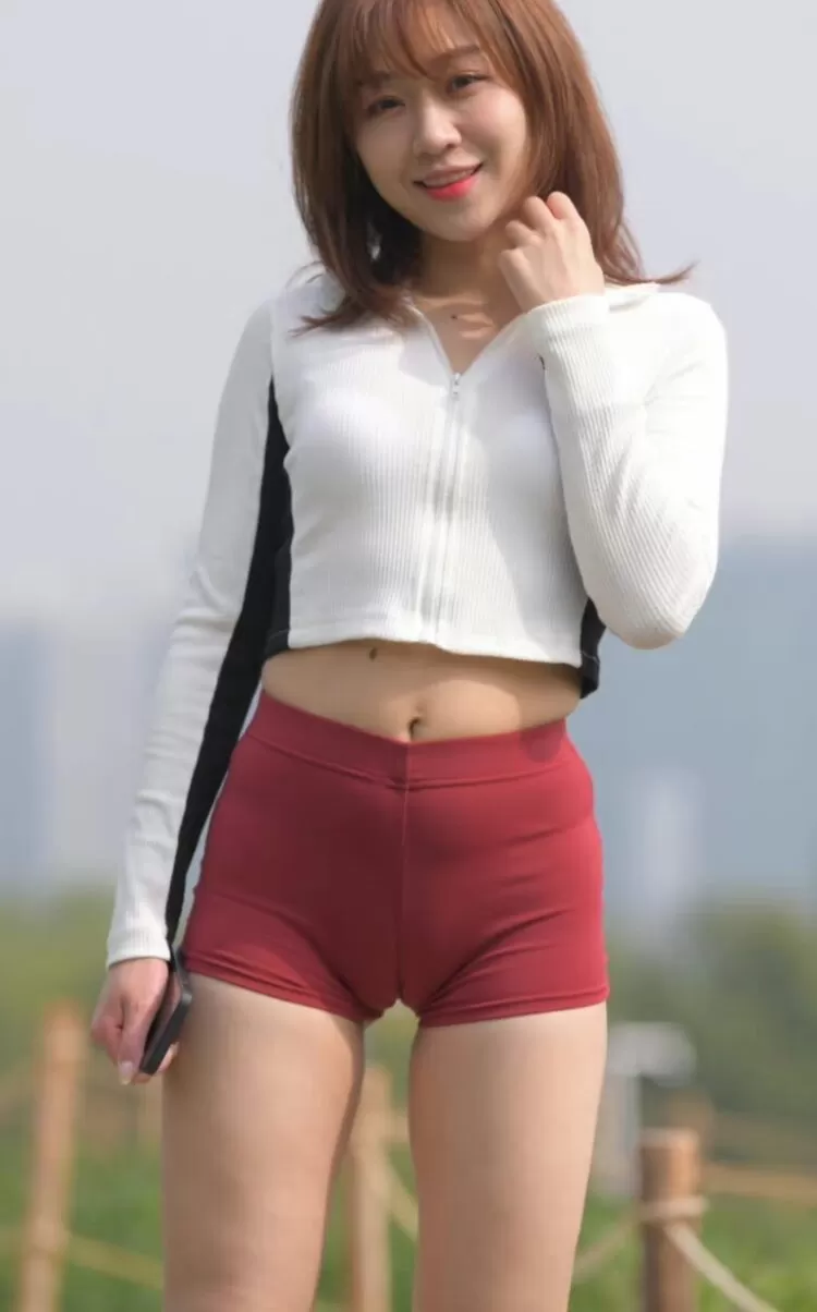 屋外で立ち姿の女性が赤い競技用ショートパンツを着用し、マンスジが際立つ股間を見せている様子