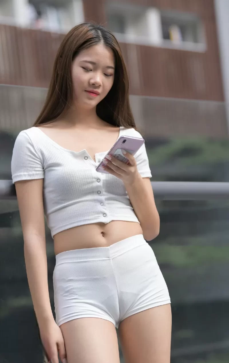 白い競技用ショートパンツを履いた女性が立ち姿勢でスマホを操作しており、タイトなパンツ越しに股間のマンスジがくっきり見える状態