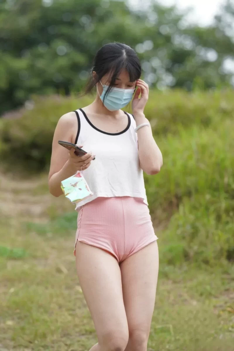 屋外でスマホを操作しながら立つ白タンクトップとピンク競技用ショートパンツの女の股間にマンスジが浮き出ている様子