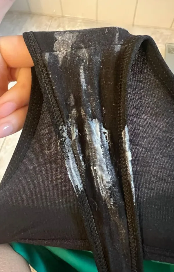 黒いパンティの股間部分に白濁したオリモノがクリーミーに付着している様子を、緑色のズボンを穿いた女性が手に持って見せているシーン