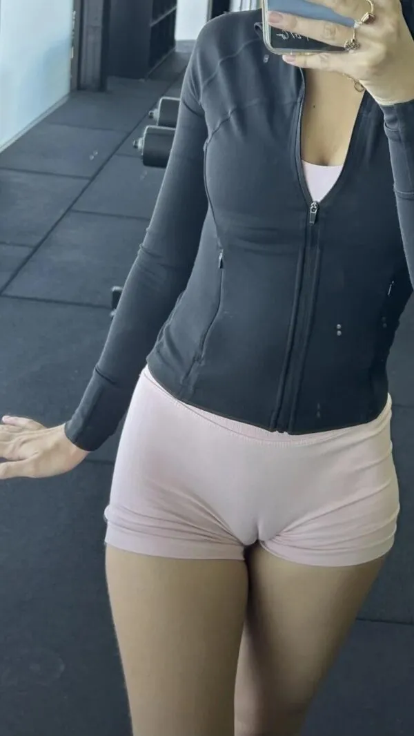 ジムの鏡前で黒いトップスにピンクのタイトなショートパンツを履いた女が、スマホで自撮りしながら前屈み気味に立ち、肉感的な股間に薄く浮かぶマンスジが卑猥に際立っているシーン。