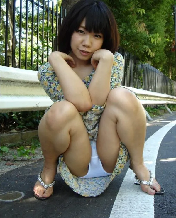 野外の道路脇で花柄ワンピースを着た女がしゃがみ込み、スカートをめくり上げて白いパンティの股間をドスケベに撮影したエロ画像